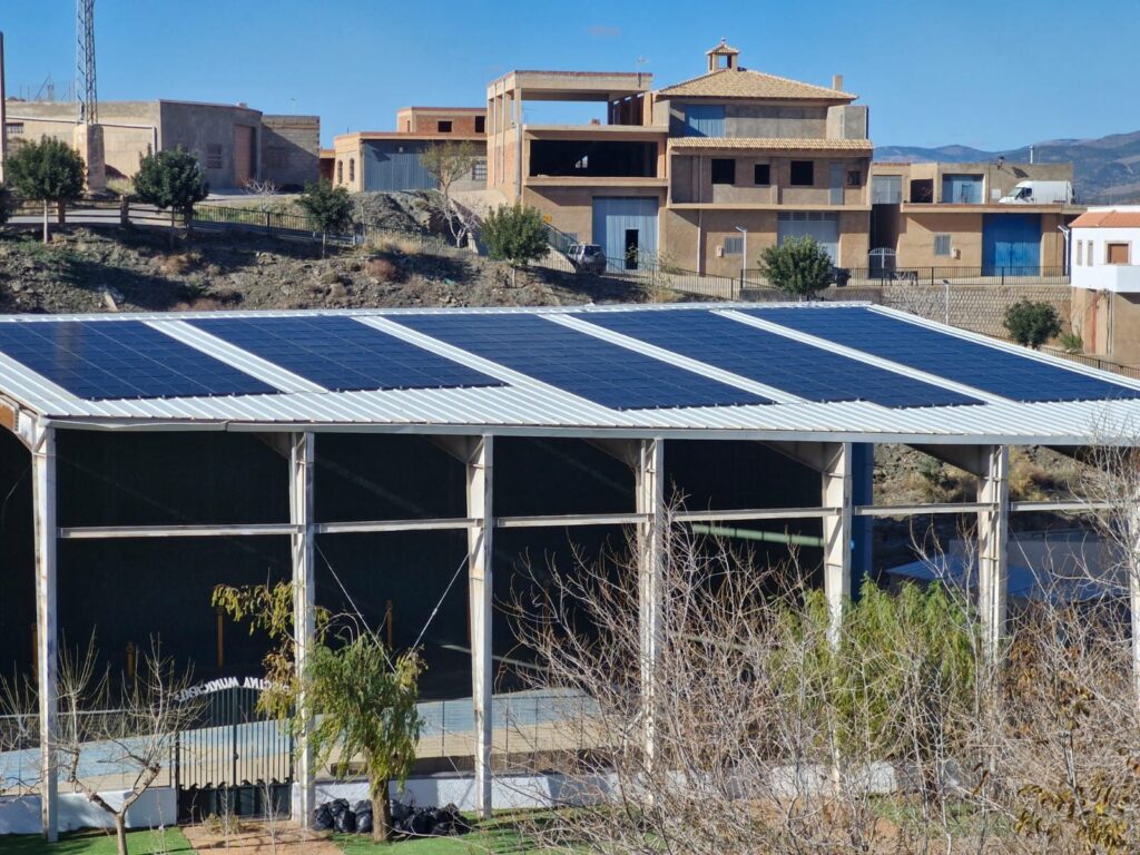 Cubierta fotovoltaica sobre la estructura de un frontón municipal con el pueblo de montaña al fondo Cubierta fotovoltaica sobre la estructura de un frontón municipal con el pueblo de montaña al fondo